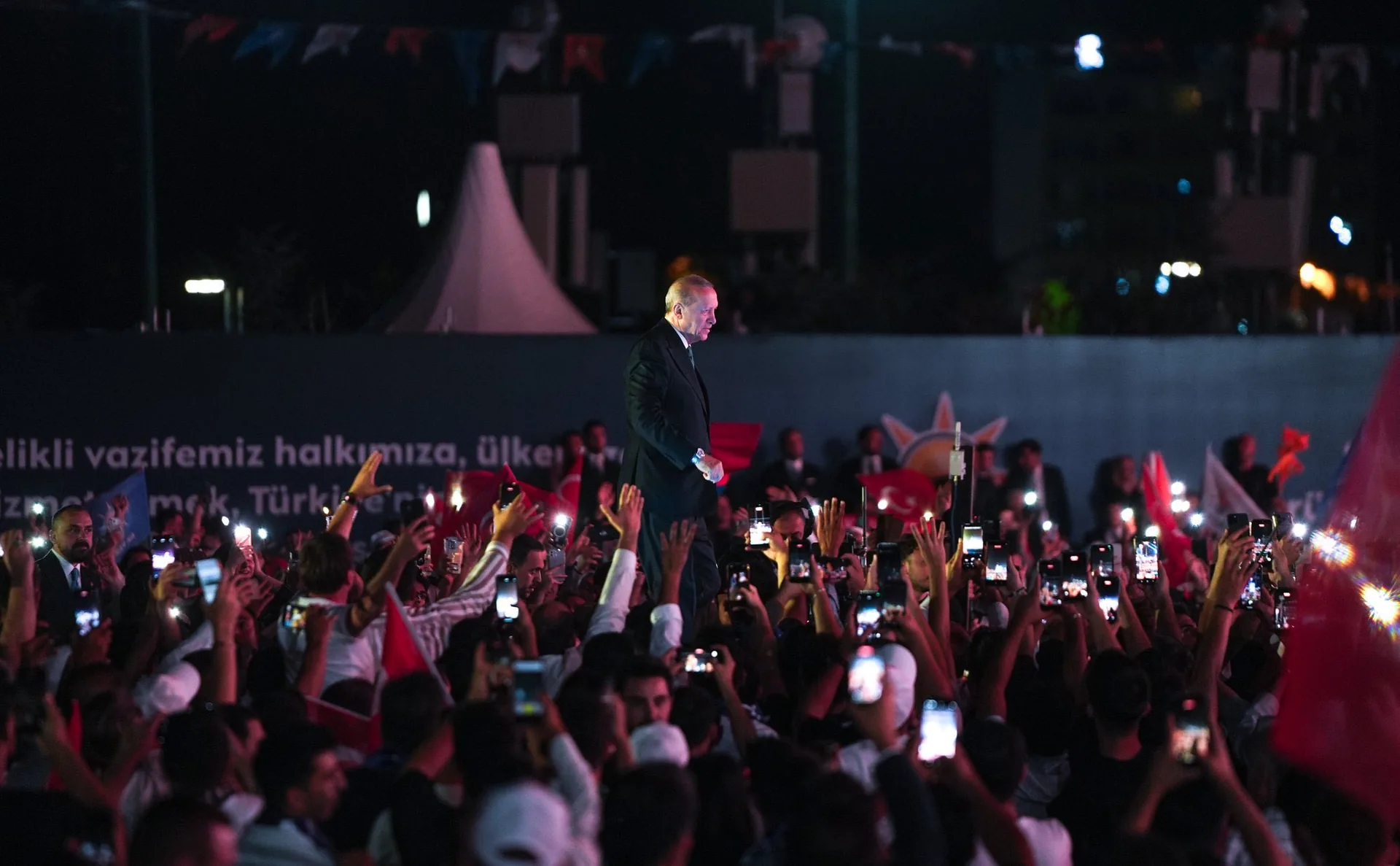 Başkent Millet Bahçesi'ndeki program alanında, telefon ışıkları ve Türk bayraklarıyla dolu kalabalığın arasında sahnede yürüyen Cumhurbaşkanı Recep Tayyip Erdoğan.